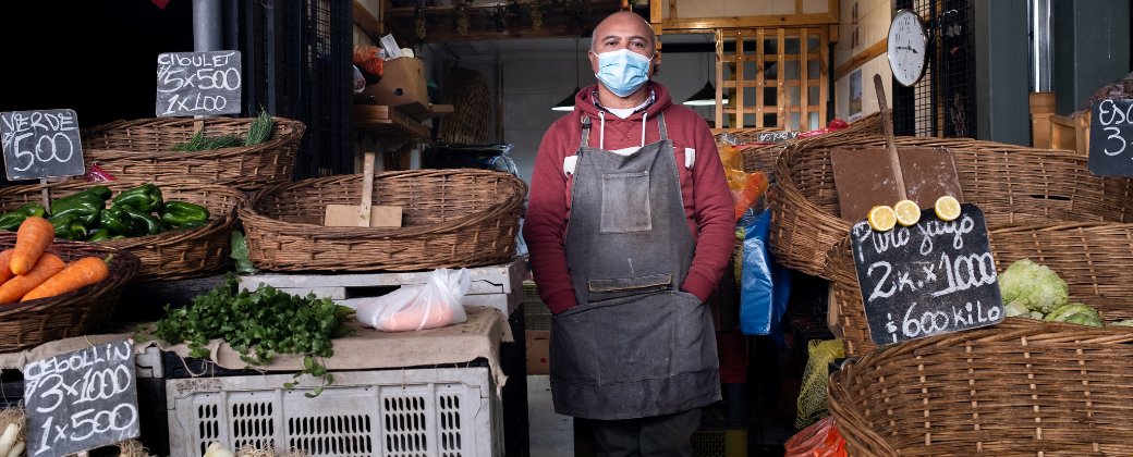 Un verdulero con barbijo posa entre los alimentos y sus precios. Créditos: Michel Garro / FAO