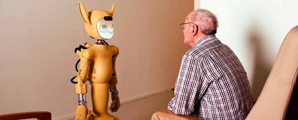 Un adulto mayor conversa frente a frente con un robot social, una escena que resume el avance de la inteligencia artificial en el acompañamiento y el cuidado. Crédito: Unsplash.